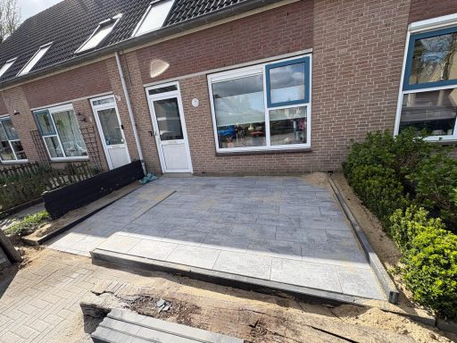 Sierbestrating voortuin Tegels liggen klaar voor een oprit bij een woonhuis, met een tuin aan de zijkant.