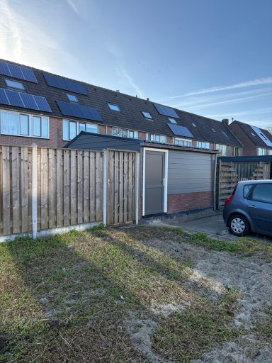 Onderhoudsarme overkapping Een parkeerplaats met een schutting en een garage, gelegen bij een rij huizen met zonnepanelen.